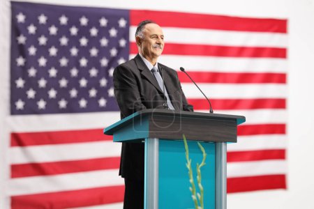 Téléchargez les photos : Politicien américain prononçant un discours sur un podium et souriant avec drapeau en arrière-plan - en image libre de droit