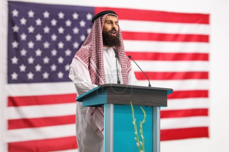 Foto de Hombre árabe dando un discurso en un pedestal frente a la bandera de EE.UU. - Imagen libre de derechos