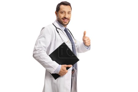 Téléchargez les photos : Jeune homme médecin gestualisant pouces vers le haut et tenant un presse-papiers isolé sur fond blanc - en image libre de droit