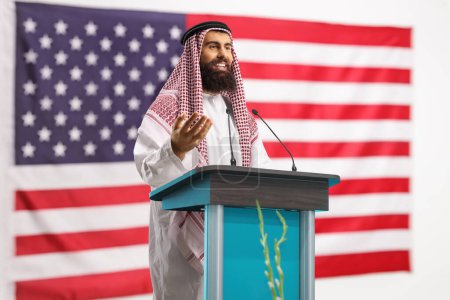 Foto de Hombre árabe dando un discurso en un pedestal y haciendo un gesto con la mano con la bandera de EE.UU. en el fondo - Imagen libre de derechos