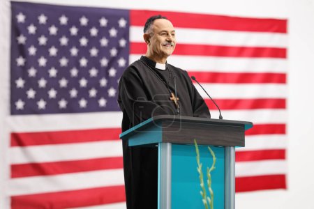 Téléchargez les photos : Prêtre souriant et debout sur un podium de discours devant un drapeau des États-Unis - en image libre de droit