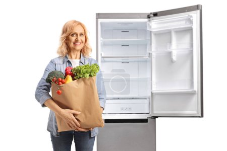 Photo for Woman with a grocery bag posing in front of an open empty fridge isolated on white background - Royalty Free Image