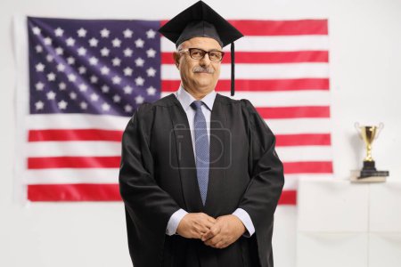 Photo for Mature man wearing a graduation gown and standing in front of a USA flag isolated on white background - Royalty Free Image