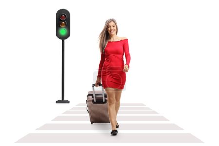 Photo for Full length portrait of a young woman walking towards camera and pulling a suitcase at a pedestrian crossing isolated on white background - Royalty Free Image