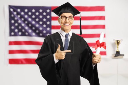 Photo for Senior man in a graduation gown holding a diploma and pointing in front of the USA flag - Royalty Free Image