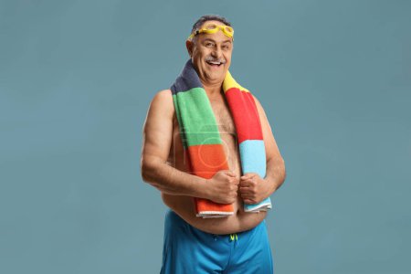 Téléchargez les photos : Gai mature l'homme en maillot de bain portant une serviette autour du cou isolé sur fond bleu - en image libre de droit