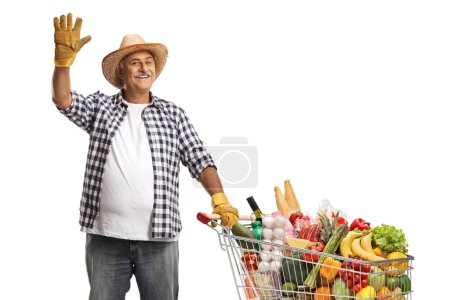 Foto de Agricultor con un carrito de compras lleno de productos alimenticios saludando a la cámara aislado sobre fondo blanco - Imagen libre de derechos