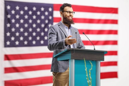 Photo for Bearded man giving a speech on a podium in front of an american flag - Royalty Free Image