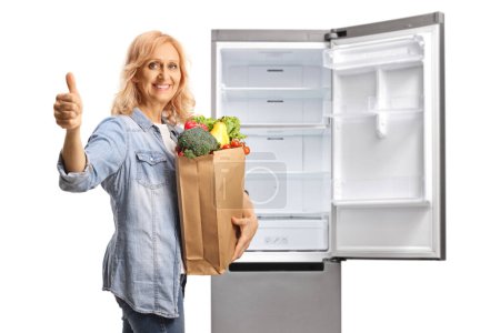 Téléchargez les photos : Femme mûre avec un sac d'épicerie devant un réfrigérateur montrant un pouce vers le haut signe isolé sur fond blanc - en image libre de droit
