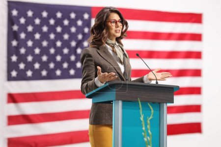 Photo for Young woman on a pedestal giving a speech and gesturing with hands with the USA flag in the background - Royalty Free Image