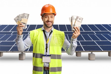 Foto de Ingeniero con chaleco de seguridad y hardhat sosteniendo montones de dinero frente a una granja fotovoltaica aislada sobre fondo blanco - Imagen libre de derechos