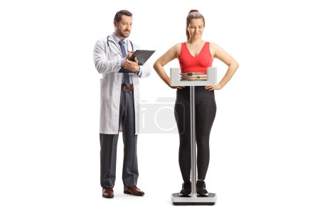 Téléchargez les photos : Jeune femme en tenue de sport pesant sur une balance médicale et médecin rédigeant un document isolé sur fond blanc - en image libre de droit