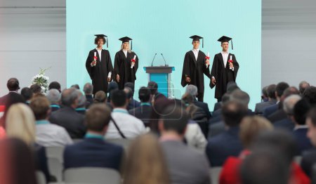 Foto de Estudiantes graduados en posesión de certificados de pie en un podio frente a la gente en la audiencia - Imagen libre de derechos