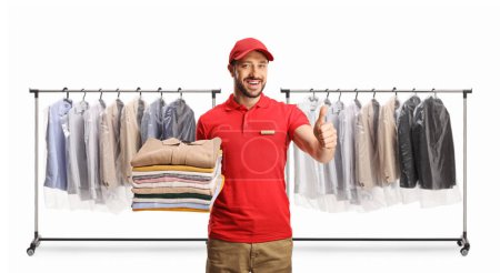 Photo for Laundry worker holding a pile of folded clothes and gesturing thumbs up in front of clothing racks isolated on a white backgroun - Royalty Free Image