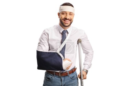 Téléchargez les photos : Jeune homme avec un bras cassé et bandage sur la tête appuyé sur une béquille et souriant isolé sur fond blanc - en image libre de droit