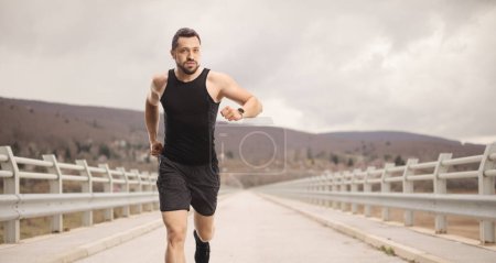 Téléchargez les photos : S'adapter jeune homme avec une smartwatch courir par temps nuageux sur un pont dans la campagne - en image libre de droit