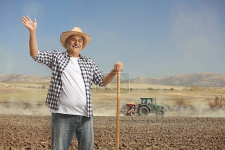 Téléchargez les photos : Fermier mature avec une pelle debout sur un champ avec un tracteur labourant le sol - en image libre de droit