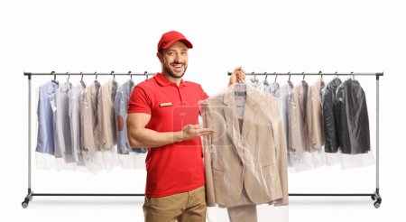 Téléchargez les photos : Travailleur masculin montrant un costume aux nettoyeurs à sec devant des rayonnages avec des vêtements suspendus isolés sur un dosseret blanc - en image libre de droit