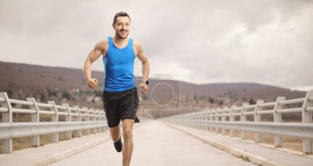 Photo for Fit man in sportswear running across a bridge on a cloudy day - Royalty Free Image