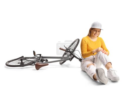 Téléchargez les photos : Jeune femme avec un vélo assis sur le sol et tenant son genou douloureux isolé sur fond blanc - en image libre de droit