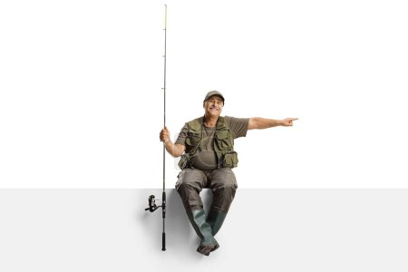 Foto de Alegre pescador maduro con una caña de pescar sentada en un panel en blanco y señalando el lado aislado sobre fondo blanco - Imagen libre de derechos