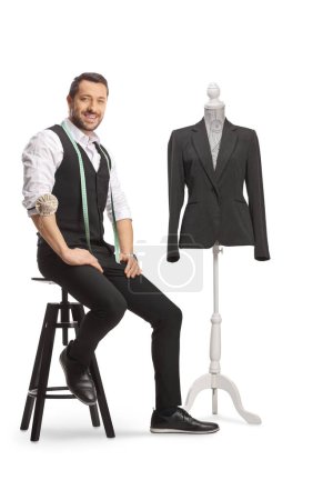 Photo for Young tailor with a measuring tape smiling at camera and sitting next to a mannequin doll with a suit isolated on white background - Royalty Free Image