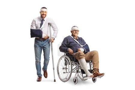 Téléchargez les photos : Jeune homme avec un bras cassé et bandage sur la tête appuyé sur une béquille et un homme mature dans un fauteuil roulant isolé sur fond blanc - en image libre de droit