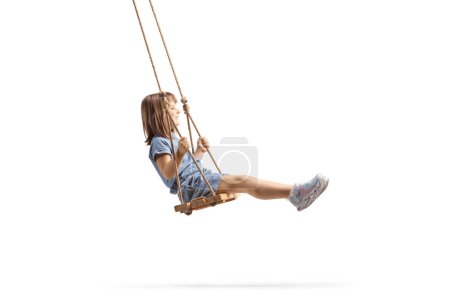 Téléchargez les photos : Plan de profil pleine longueur d'une heureuse petite fille se balançant sur une balançoire en bois isolée sur fond blanc - en image libre de droit