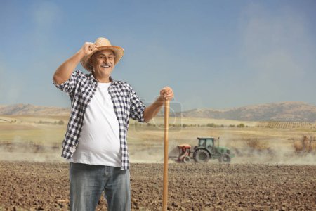 Téléchargez les photos : Portrait d'un fermier mature heureux avec une pelle debout sur un champ poussiéreux avec tracteur - en image libre de droit