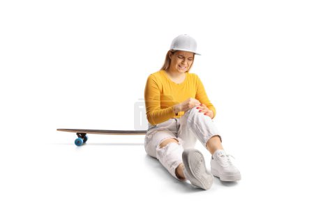 Photo for Young female with a skateboard sitting on the floor and holding her injured knee isolated on white background - Royalty Free Image