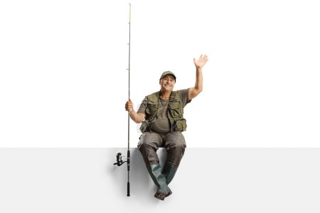 Foto de Pescador maduro con una caña de pescar sentado en un panel en blanco y saludando aislado sobre fondo blanco - Imagen libre de derechos
