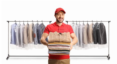 Photo for Male worker holding a pile of folded clothes in front of clothing racks isolated on a white background - Royalty Free Image