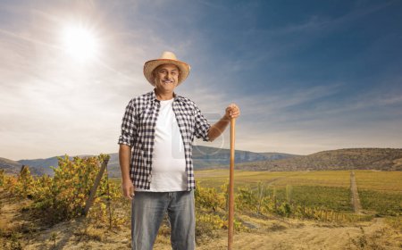 Foto de Granjero maduro con una pala de pie en un campo y sonriendo - Imagen libre de derechos