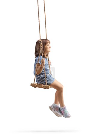 Téléchargez les photos : Plan de profil pleine longueur d'une heureuse petite fille assise sur une balançoire en bois isolée sur fond blanc - en image libre de droit