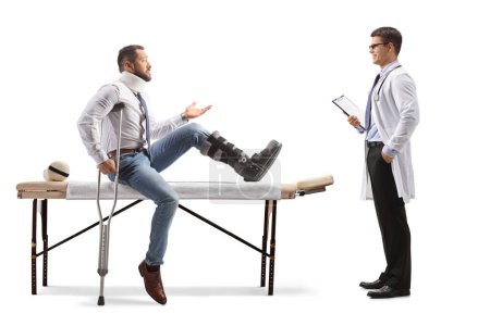 Téléchargez les photos : Homme blessé avec une botte orthopédique et un col assis sur une table de thérapie et parlant à un médecin isolé sur fond blanc - en image libre de droit