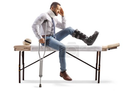 Téléchargez les photos : Homme blessé avec une botte orthopédique et un collier cervical assis sur une table de thérapie et tenant sa tête isolée sur fond blanc - en image libre de droit