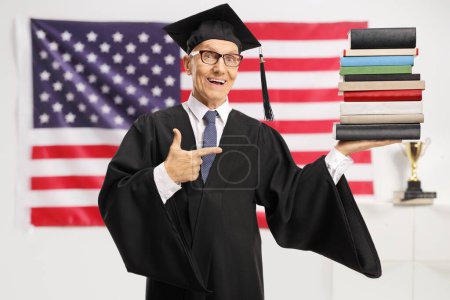 Téléchargez les photos : Homme aîné portant une robe de remise des diplômes et tenant une pile de livres devant le drapeau des États-Unis - en image libre de droit