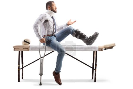 Téléchargez les photos : Homme blessé avec une botte orthopédique et un col assis sur une table de thérapie et faisant un geste avec la main isolée sur fond blanc - en image libre de droit