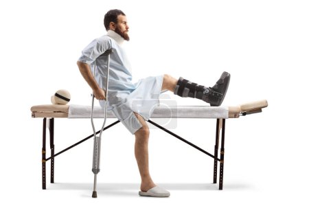 Foto de Hombre barbudo con una bata de hospital sentado en una mesa médica con una pierna y cuello rotos aislados sobre fondo blanco - Imagen libre de derechos