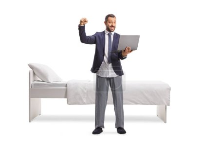 Foto de Hombre en traje y pijama de fondo usando un ordenador portátil y haciendo gestos de felicidad frente a una cama aislada sobre fondo blanco - Imagen libre de derechos