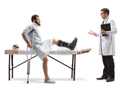 Téléchargez les photos : Homme blessé avec une botte orthopédique et un col assis sur une table de thérapie et ayant une conversation avec un médecin isolé sur fond blanc - en image libre de droit