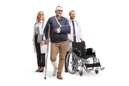 Téléchargez les photos : Médecins avec un fauteuil roulant debout derrière un patient masculin mature avec une jambe et un bras cassés isolés sur fond blanc - en image libre de droit