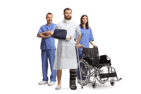 Foto de Paciente con una bata de hospital con una pierna y un brazo rotos de pie con un equipo de trabajadores médicos aislados en el fondo blanco - Imagen libre de derechos