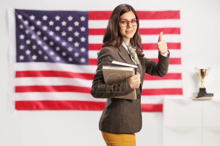 Foto de Joven hembra con gafas sosteniendo libros frente a la bandera de USA y haciendo gestos de pulgares u - Imagen libre de derechos