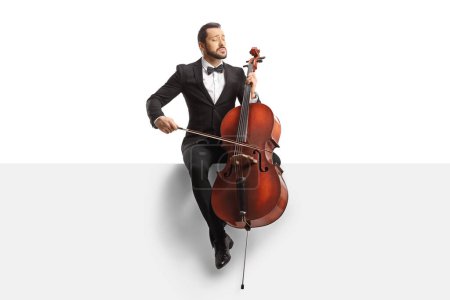 Photo for Male musician in a black suit and bow-tie sitting on a blank panel with a cello isolated on white background - Royalty Free Image