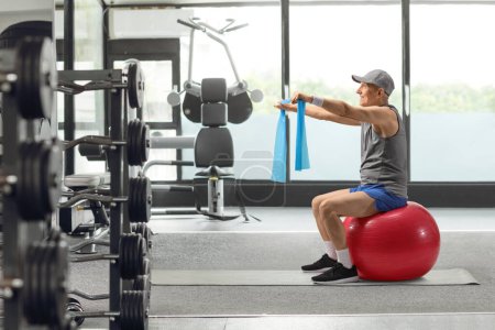 Photo for Elderly man in sportswear sitting on a fitness ball and exercising with an elastic band at the gym - Royalty Free Image