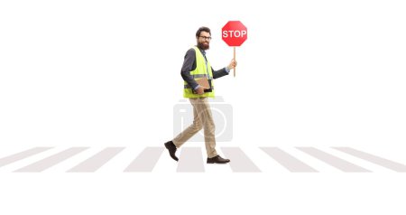 Téléchargez les photos : Plan complet d'un homme barbu dans un gilet de sécurité marchant avec un panneau d'arrêt à un passage à niveau pour piétons isolé sur fond blanc - en image libre de droit