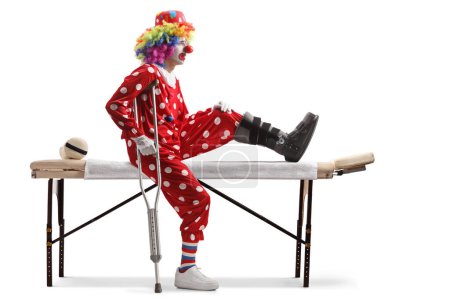Téléchargez les photos : Clown blessé avec une attelle de marche assise sur un lit thérapeutique isolé sur fond blanc - en image libre de droit