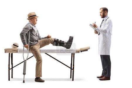 Téléchargez les photos : Médecin rédigeant une ordonnance et un patient âgé assis sur un lit de physiothérapie avec une attelle sur sa jambe isolée sur fond blanc - en image libre de droit