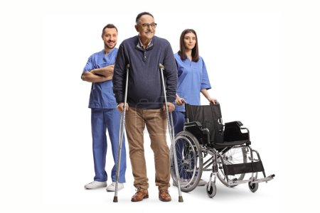 Foto de Hombre maduro con muletas parado frente a trabajadores de la salud con una silla de ruedas aislada sobre fondo blanco - Imagen libre de derechos